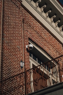 A red brick building facade featuring a distinctive white cornice at the top. A surveillance camera is mounted above a large wooden door, which is flanked by a vintage-style exterior light fixture. An ornate railing runs horizontally across the lower part of the image.