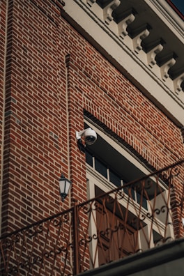 A red brick building facade featuring a distinctive white cornice at the top. A surveillance camera is mounted above a large wooden door, which is flanked by a vintage-style exterior light fixture. An ornate railing runs horizontally across the lower part of the image.