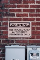 A metal sign mounted on a red brick wall indicates a restricted area for authorized personnel only. Below the sign, there are two utility boxes, one of which has a small warning label indicating high voltage.