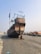 A large wooden boat with a flagged mast is situated on a sandy surface near a stone embankment. The boat has a distinctively classic design with intricate railings and is surrounded by a few concrete blocks. The sky is clear with a light blue hue.