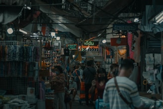 A bustling market scene with people walking through a narrow aisle surrounded by various small shops. The area is covered by a canopy, and numerous hanging lights illuminate the space. Displays of colorful fabrics and trinkets can be seen, along with signs written in a non-English language. The atmosphere suggests a busy and crowded environment.