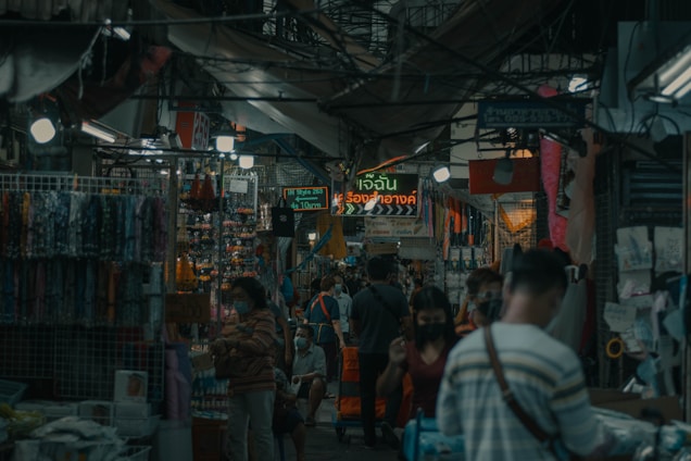 A bustling market scene with people walking through a narrow aisle surrounded by various small shops. The area is covered by a canopy, and numerous hanging lights illuminate the space. Displays of colorful fabrics and trinkets can be seen, along with signs written in a non-English language. The atmosphere suggests a busy and crowded environment.