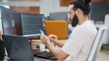 A person with a beard is seated at an office desk, looking at a smartphone. Two large computer monitors display code and digital content. A Dell laptop is also on the desk, along with a cup and a keyboard.
