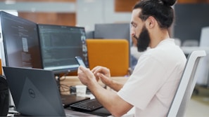 A person with a beard is seated at an office desk, looking at a smartphone. Two large computer monitors display code and digital content. A Dell laptop is also on the desk, along with a cup and a keyboard.