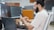 A person with a beard is seated at an office desk, looking at a smartphone. Two large computer monitors display code and digital content. A Dell laptop is also on the desk, along with a cup and a keyboard.