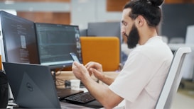A person with a beard is seated at an office desk, looking at a smartphone. Two large computer monitors display code and digital content. A Dell laptop is also on the desk, along with a cup and a keyboard.