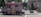 A pink ice cream truck with various images of ice cream cones and waffles parked on the street. Several people are in the background, including someone wearing an inflatable dinosaur costume near a building entrance. The environment appears urban, with trees and buildings visible and a person pushing a stroller.