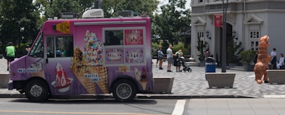 A pink ice cream truck with various images of ice cream cones and waffles parked on the street. Several people are in the background, including someone wearing an inflatable dinosaur costume near a building entrance. The environment appears urban, with trees and buildings visible and a person pushing a stroller.