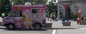 A pink ice cream truck with various images of ice cream cones and waffles parked on the street. Several people are in the background, including someone wearing an inflatable dinosaur costume near a building entrance. The environment appears urban, with trees and buildings visible and a person pushing a stroller.