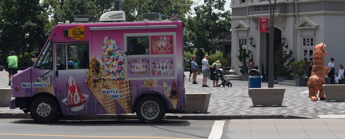 A pink ice cream truck with various images of ice cream cones and waffles parked on the street. Several people are in the background, including someone wearing an inflatable dinosaur costume near a building entrance. The environment appears urban, with trees and buildings visible and a person pushing a stroller.
