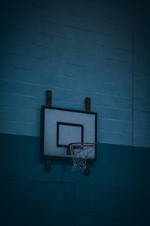 A basketball hoop is mounted on a blue gymnasium wall, featuring a white backboard with a black square target painted in the center. The red hoop has a white net attached.