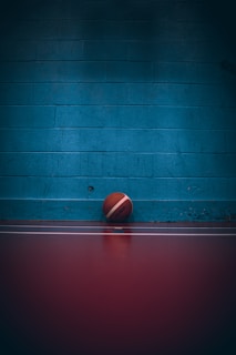 A basketball is resting against a blue brick wall on a dark red-colored floor. The lighting is dim, casting a shadow over the wall and the ball.
