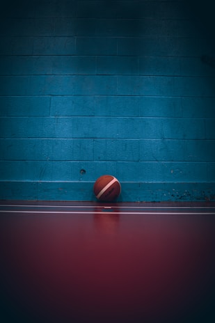A basketball is resting against a blue brick wall on a dark red-colored floor. The lighting is dim, casting a shadow over the wall and the ball.