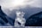An intricately carved ice sculpture of an animal, possibly a reindeer, stands in the foreground against a backdrop of misty mountains and a cloudy sky. The ice is translucent and reflects the ambient light, adding a mystical quality.
