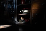 A wooden cabinet with glass doors contains neatly arranged glassware. An open shelf reveals ceramic dishes in muted lighting, creating a play of shadows and highlights.