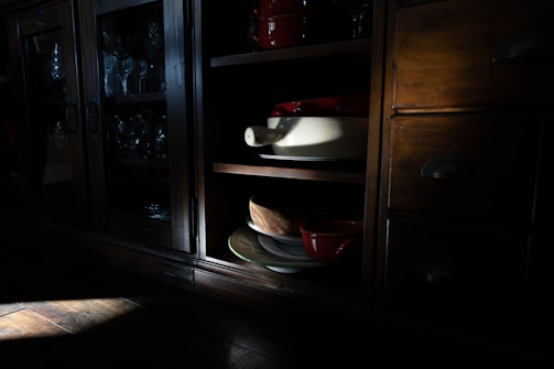 A wooden cabinet with glass doors contains neatly arranged glassware. An open shelf reveals ceramic dishes in muted lighting, creating a play of shadows and highlights.