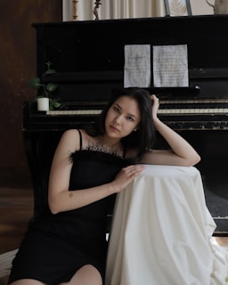 A woman in a black dress is sitting beside a piano, resting her head on one hand while leaning on a draped white cloth. The piano has sheet music on the stand and some decorative elements, including a small plant and candle.
