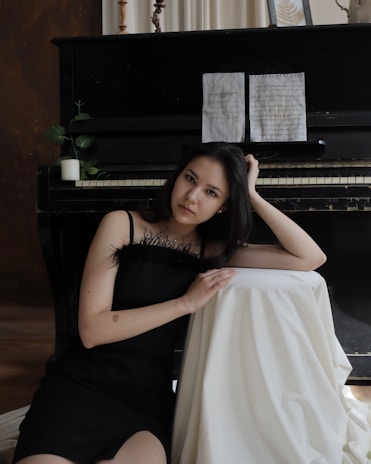 A woman in a black dress is sitting beside a piano, resting her head on one hand while leaning on a draped white cloth. The piano has sheet music on the stand and some decorative elements, including a small plant and candle.