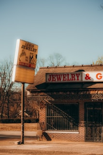 A vintage jewelry store with a large sign reading 'Lewis Jewelry' stands by an empty street. The building is constructed of brick and features a sign that reads 'JEWELRY & G...' The surroundings include bare trees under a clear, bright blue sky, suggesting a cold season.