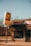 A vintage jewelry store with a large sign reading 'Lewis Jewelry' stands by an empty street. The building is constructed of brick and features a sign that reads 'JEWELRY & G...' The surroundings include bare trees under a clear, bright blue sky, suggesting a cold season.
