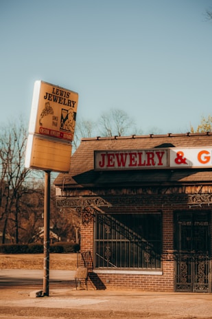 A vintage jewelry store with a large sign reading 'Lewis Jewelry' stands by an empty street. The building is constructed of brick and features a sign that reads 'JEWELRY & G...' The surroundings include bare trees under a clear, bright blue sky, suggesting a cold season.
