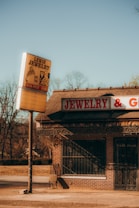 A vintage jewelry store with a large sign reading 'Lewis Jewelry' stands by an empty street. The building is constructed of brick and features a sign that reads 'JEWELRY & G...' The surroundings include bare trees under a clear, bright blue sky, suggesting a cold season.