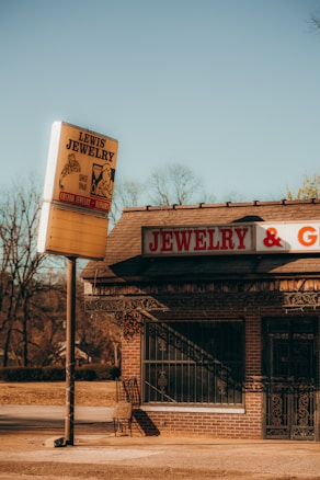 A vintage jewelry store with a large sign reading 'Lewis Jewelry' stands by an empty street. The building is constructed of brick and features a sign that reads 'JEWELRY & G...' The surroundings include bare trees under a clear, bright blue sky, suggesting a cold season.