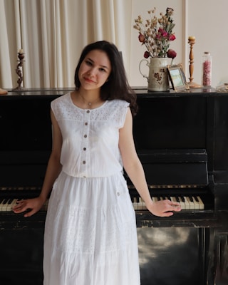 A person wearing a white dress stands in front of a black upright piano. On top of the piano are decorative items including a candle holder, a pitcher with dried flowers, and a framed picture. The background features light-colored curtains.