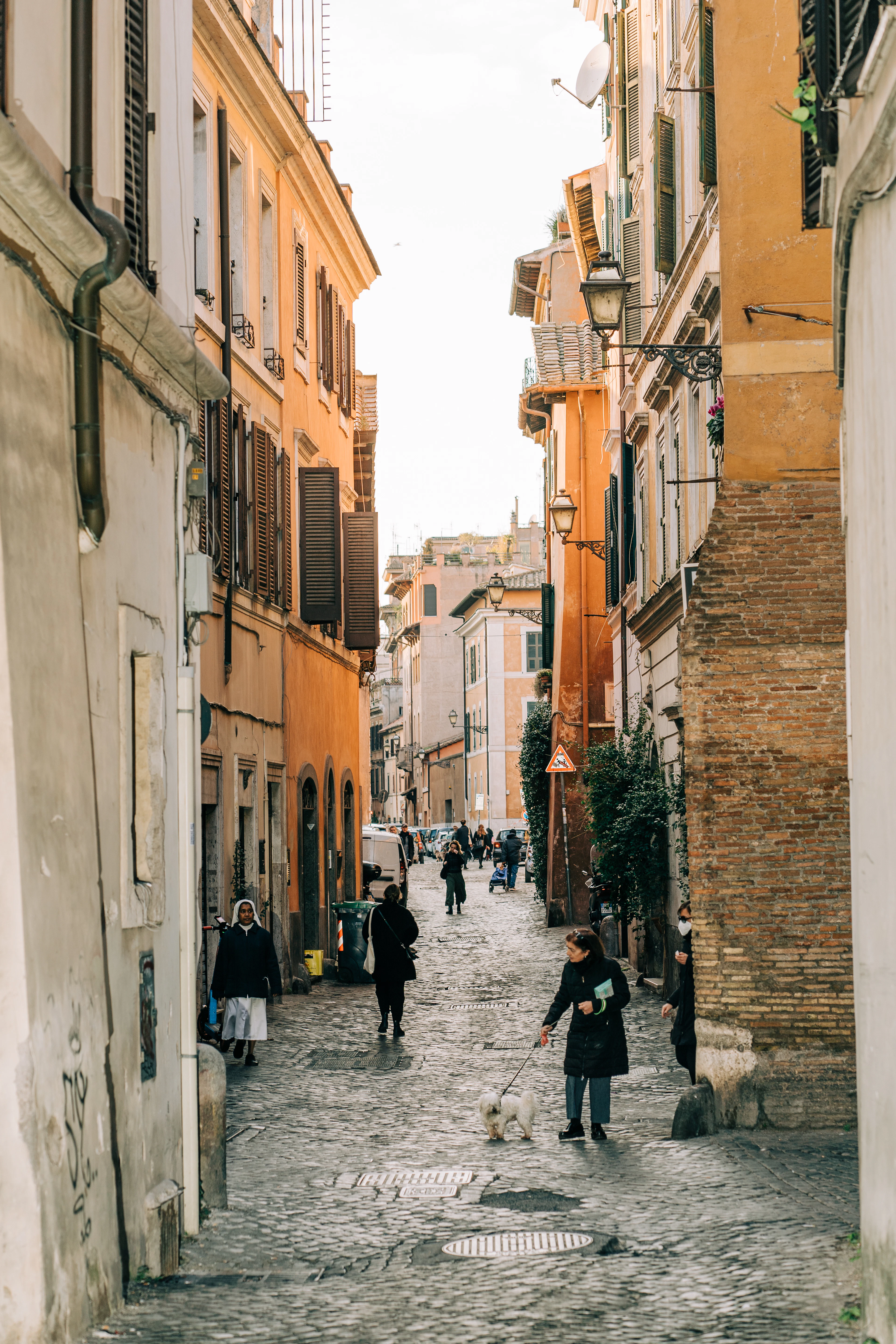 A narrow cobbled lane in Trastevere, Rome, lined with ochre and cream buildings, shuttered windows and strolling residents