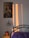 A bedroom scene featuring a framed artwork of a person in a colorful headdress on the wall. Warm sunlight casts long rectangles of light and shadow on the wall. A small potted plant sits on a wooden bedside table next to a bed with a metal headboard and a purple bedspread.