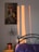 A bedroom scene featuring a framed artwork of a person in a colorful headdress on the wall. Warm sunlight casts long rectangles of light and shadow on the wall. A small potted plant sits on a wooden bedside table next to a bed with a metal headboard and a purple bedspread.
