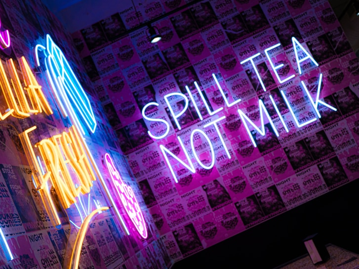 Bright neon signs in blue and yellow with a mix of posters create an energetic and vibrant atmosphere. The phrases 'SPILL TEA NOT MILK' and another partially visible sign stand out against a backdrop of various pink and purple posters.