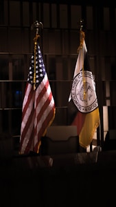 Two flags are displayed indoors. The American flag with red, white, and blue colors is placed next to a flag representing the Seminole Tribe of Florida. Both flags are on flagpoles with gold fringes.