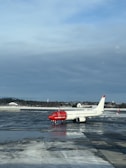 An airplane with a red nose and predominantly white body is stationed on a wet, reflective tarmac. The words 'norwegian.com' are visible on the side of the aircraft. The sky is overcast with a mix of blue and gray clouds. In the background, there is a line of trees and a few small buildings partially covered in snow.