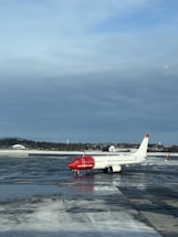 An airplane with a red nose and predominantly white body is stationed on a wet, reflective tarmac. The words 'norwegian.com' are visible on the side of the aircraft. The sky is overcast with a mix of blue and gray clouds. In the background, there is a line of trees and a few small buildings partially covered in snow.