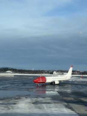 An airplane with a red nose and predominantly white body is stationed on a wet, reflective tarmac. The words 'norwegian.com' are visible on the side of the aircraft. The sky is overcast with a mix of blue and gray clouds. In the background, there is a line of trees and a few small buildings partially covered in snow.