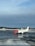 An airplane with a red nose and predominantly white body is stationed on a wet, reflective tarmac. The words 'norwegian.com' are visible on the side of the aircraft. The sky is overcast with a mix of blue and gray clouds. In the background, there is a line of trees and a few small buildings partially covered in snow.