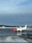 An airplane with a red nose and predominantly white body is stationed on a wet, reflective tarmac. The words 'norwegian.com' are visible on the side of the aircraft. The sky is overcast with a mix of blue and gray clouds. In the background, there is a line of trees and a few small buildings partially covered in snow.