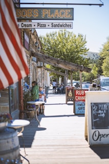 A wooden boardwalk lined with rustic shops and signs, featuring an American flag in the foreground. Several people, including a child wearing a sports jersey, walk along the boardwalk. Nearby, tables are set up outside a store with signs advertising pizza and gift shops. Trees and parked cars are visible in the background.