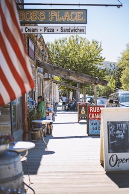 A wooden boardwalk lined with rustic shops and signs, featuring an American flag in the foreground. Several people, including a child wearing a sports jersey, walk along the boardwalk. Nearby, tables are set up outside a store with signs advertising pizza and gift shops. Trees and parked cars are visible in the background.