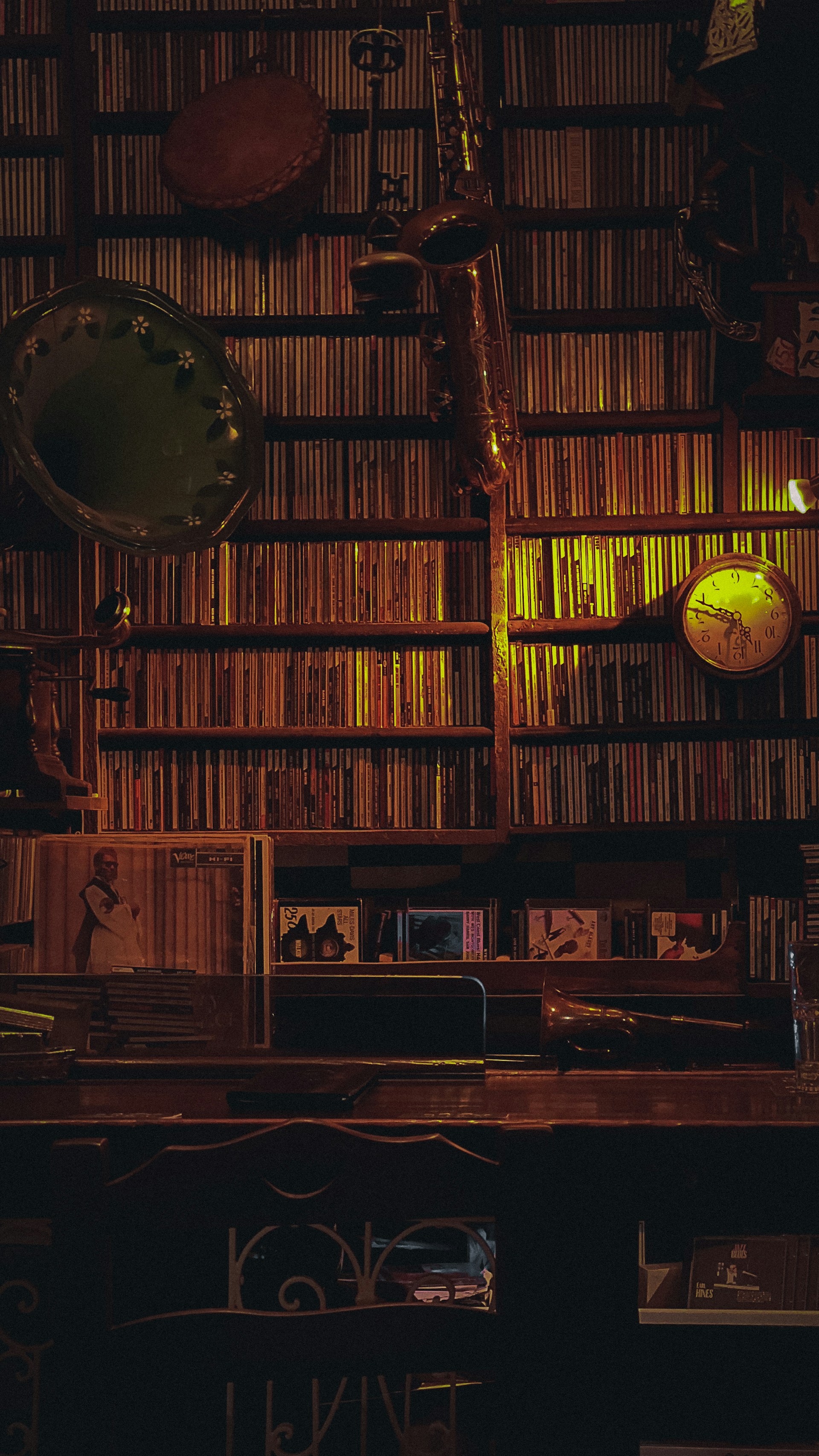A well-organized collection of CDs is arranged on wall-mounted shelves. A saxophone and a drum with soft ambient lighting highlight the musical theme. A vintage clock and a few album covers add to the atmosphere, with a warm and nostalgic ambiance.