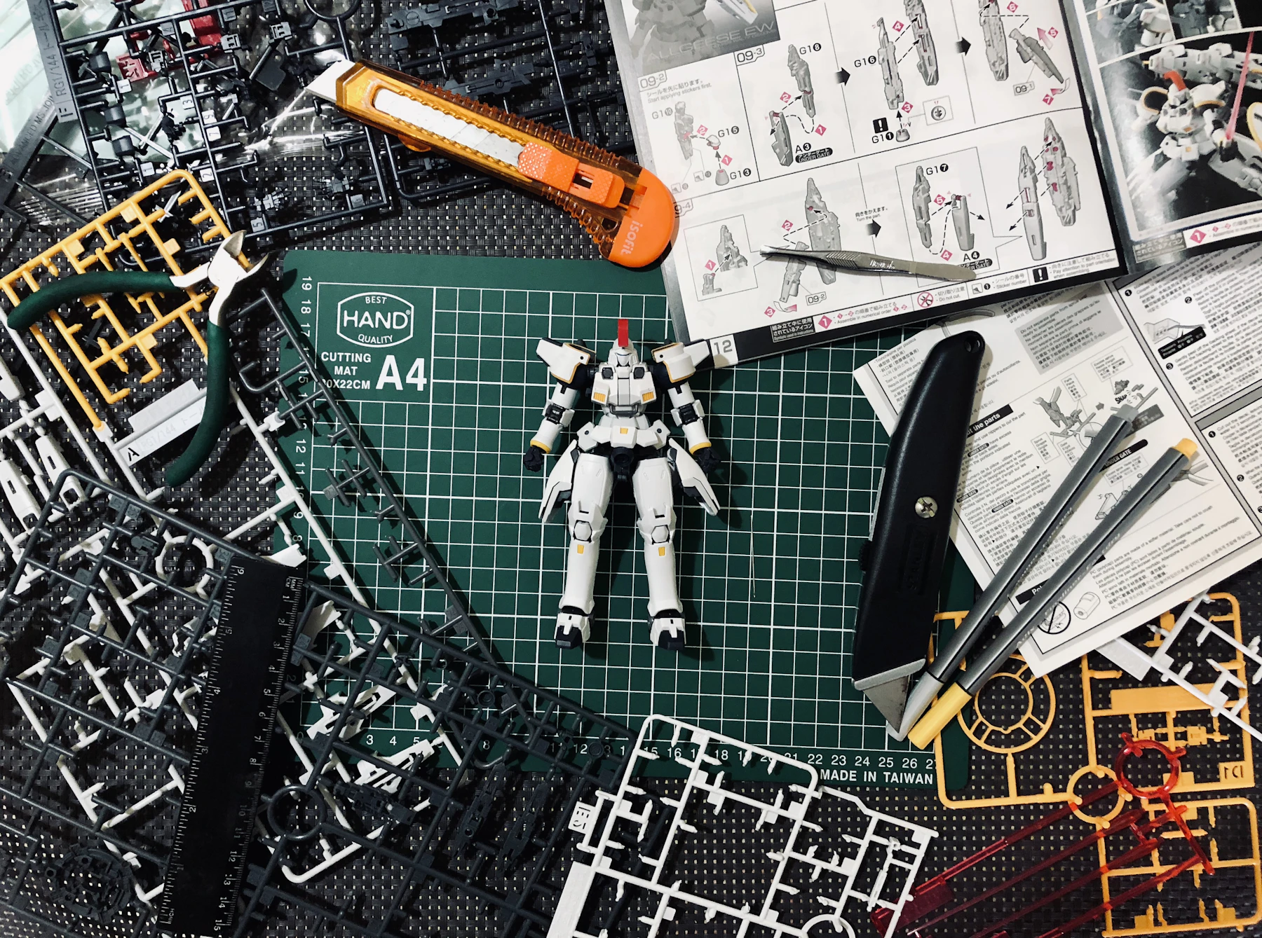 A green cutting mat covered in black, white and yellow part frames, with a small white robot model, a craft knife and instruction sheets