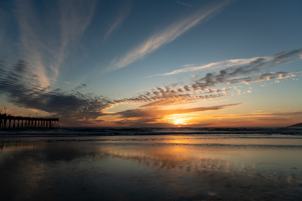 the sun is setting over a beach with a pier in the distance