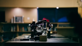 An indoor scene featuring a modern coffee brewing setup on a wooden counter. In the foreground, a glass coffee pot and various coffee-making equipment can be seen. The background is softly blurred, showing shelves with books and a warm ambiance created by soft lighting.