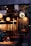 A cozy, warmly lit boutique or cafe with stylish clothes hanging on racks. There are round pendant lights and a decorative chalkboard. Floral arrangements adorn tables, and a person is seated and engaged in dining or working.