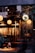 A cozy, warmly lit boutique or cafe with stylish clothes hanging on racks. There are round pendant lights and a decorative chalkboard. Floral arrangements adorn tables, and a person is seated and engaged in dining or working.