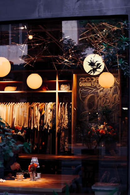 A cozy, warmly lit boutique or cafe with stylish clothes hanging on racks. There are round pendant lights and a decorative chalkboard. Floral arrangements adorn tables, and a person is seated and engaged in dining or working.