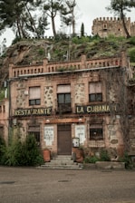 A rustic building with stone and brick facade, featuring signs indicating it is a restaurant named 'La Cubanita.' The exterior shows slightly worn elements, creating a vintage look, with plants growing from the top and sides. An old castle is visible on the hill behind it, surrounded by trees and greenery.