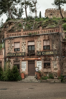 A rustic building with stone and brick facade, featuring signs indicating it is a restaurant named 'La Cubanita.' The exterior shows slightly worn elements, creating a vintage look, with plants growing from the top and sides. An old castle is visible on the hill behind it, surrounded by trees and greenery.