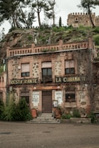 A rustic building with stone and brick facade, featuring signs indicating it is a restaurant named 'La Cubanita.' The exterior shows slightly worn elements, creating a vintage look, with plants growing from the top and sides. An old castle is visible on the hill behind it, surrounded by trees and greenery.